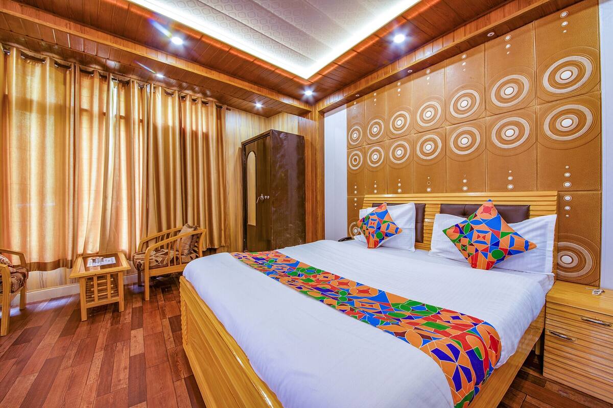 Una habitación de hotel con piso de madera, una cama grande con ropa de cama colorida, un cabecero de madera con diseños circulares, un armario de madera y una pequeña mesa con sillas.