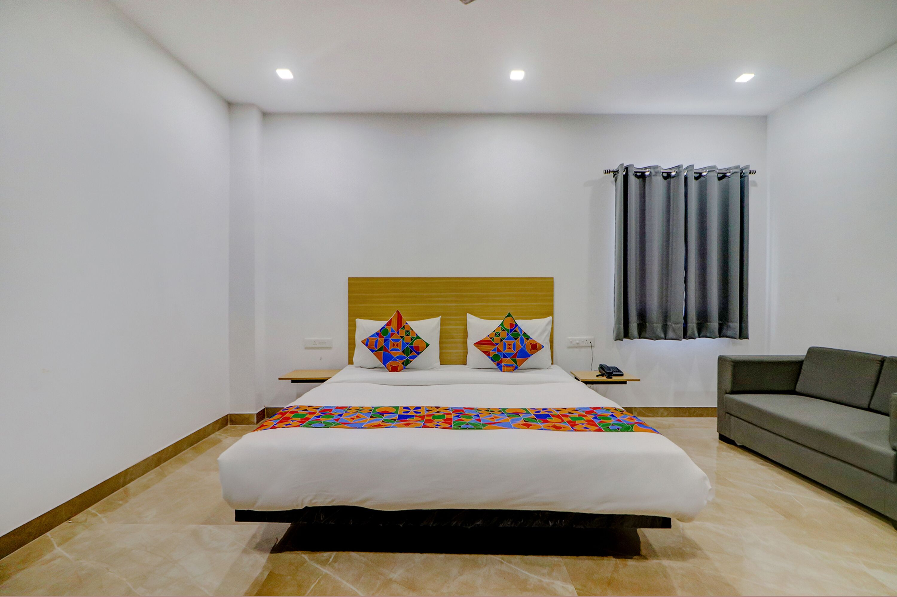 Une chambre d’hôtel avec un grand lit, deux oreillers, une table de chevet, un canapé gris et une fenêtre avec des rideaux.