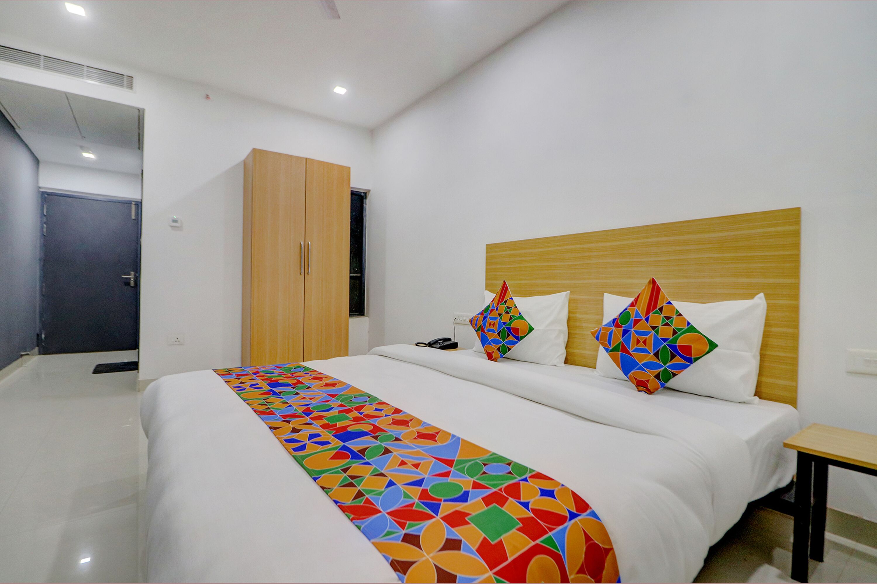 A hotel room with a bed, a wooden wardrobe, a bedside table, and a colorful bedspread.