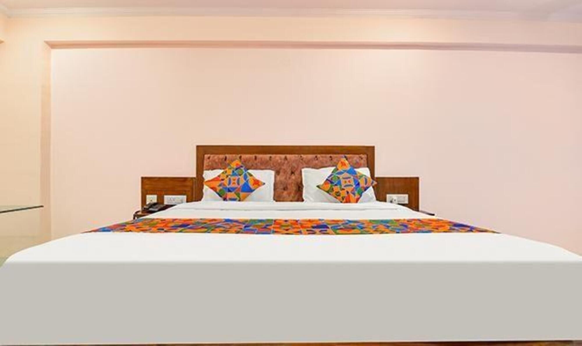 A neatly made bed with a colorful quilt and two decorative pillows, a wooden headboard, and a plain wall in a hotel room.