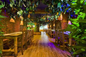 Ein Restaurant mit Holztischen und -stühlen, hängenden Laternen und einer Backsteinwand im Hintergrund.