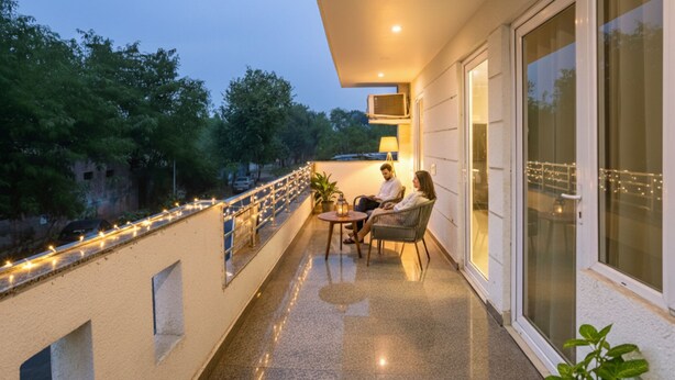 Ein Balkon mit zwei Personen, die an einem Tisch sitzen, einer Geländer mit Lichterketten sowie Blick auf Bäume und ein Gebäude im Hintergrund.