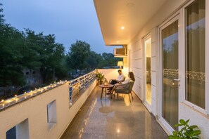 A balcony with two people seated at a table, a railing with string lights, and a view of trees and a building in the background. - FabHotel White House BNB - Nr. Yashobhoomi Convention Centre (New Delhi)