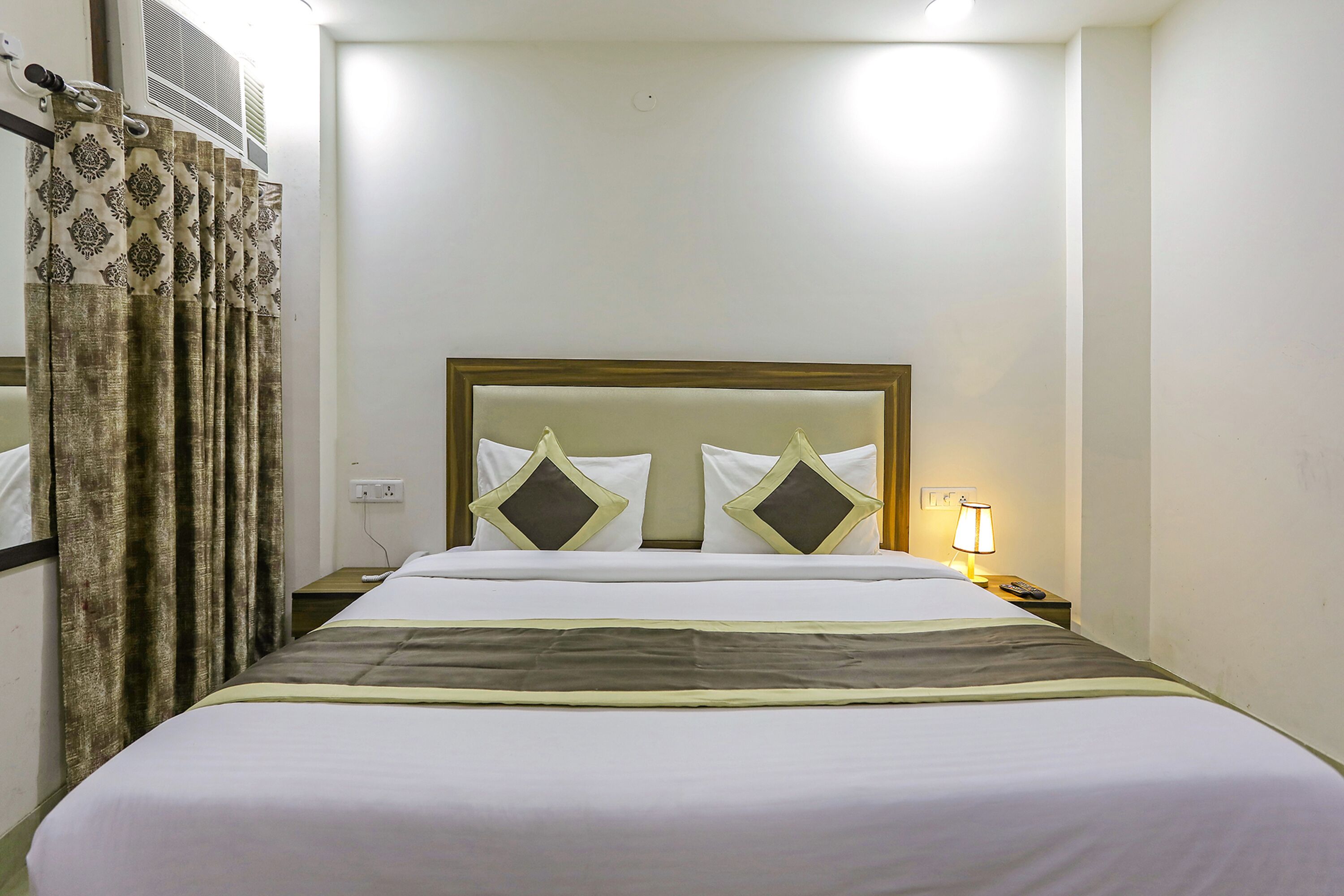 A hotel room with a bed, curtains, a nightstand with a lamp, and a wall-mounted air conditioning unit.