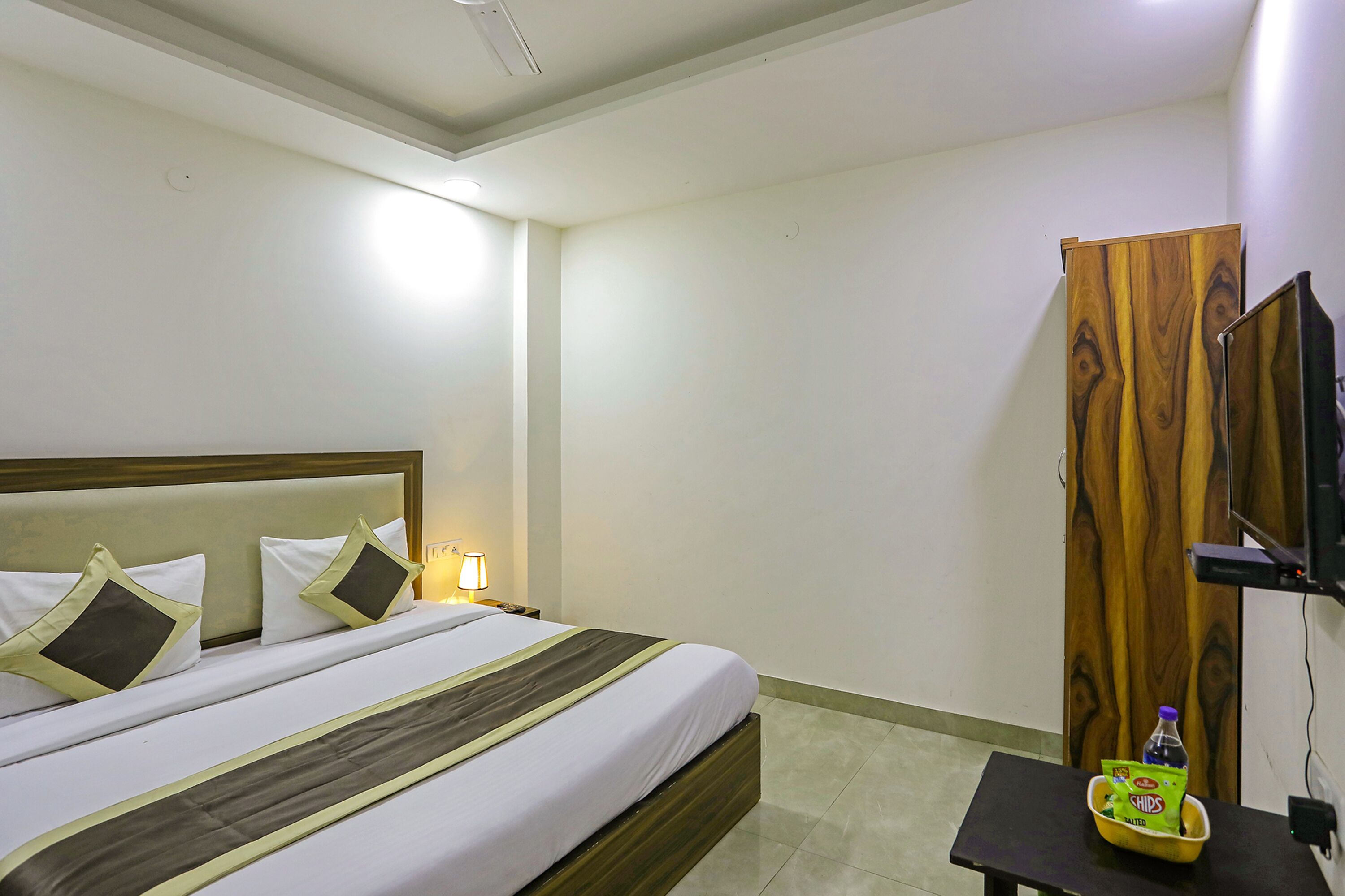 Habitación de hotel con cama, mesita de noche con lámpara, armario de madera y una pequeña mesa con aperitivos y una botella.