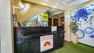 A hotel reception desk with a sign for 'Tab Hotels' and 'Amazing Inn' by SRK Groups of Hotels. There is a fan, a potted plant, and a wall with a floral mural.