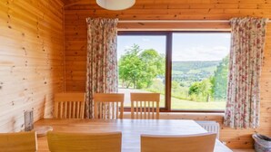 Eine Holzhütte mit Esstisch und Stühlen, großen Fenstern mit Vorhängen und Blick auf eine grüne Landschaft.