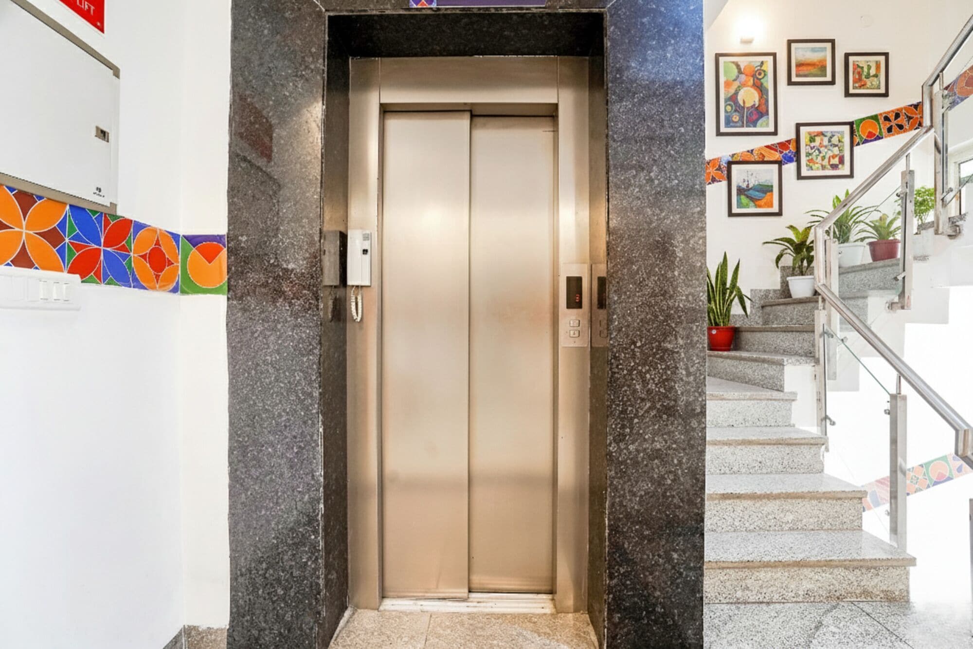 Ein Aufzug mit Bedienfeld und digitaler Anzeige, der zu einer Treppe mit Geländern und Pflanzkübeln führt.