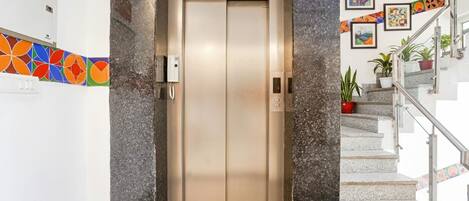 Un ascensor con panel de control y pantalla digital, que da acceso a una escalera con barandillas y plantas en macetas.