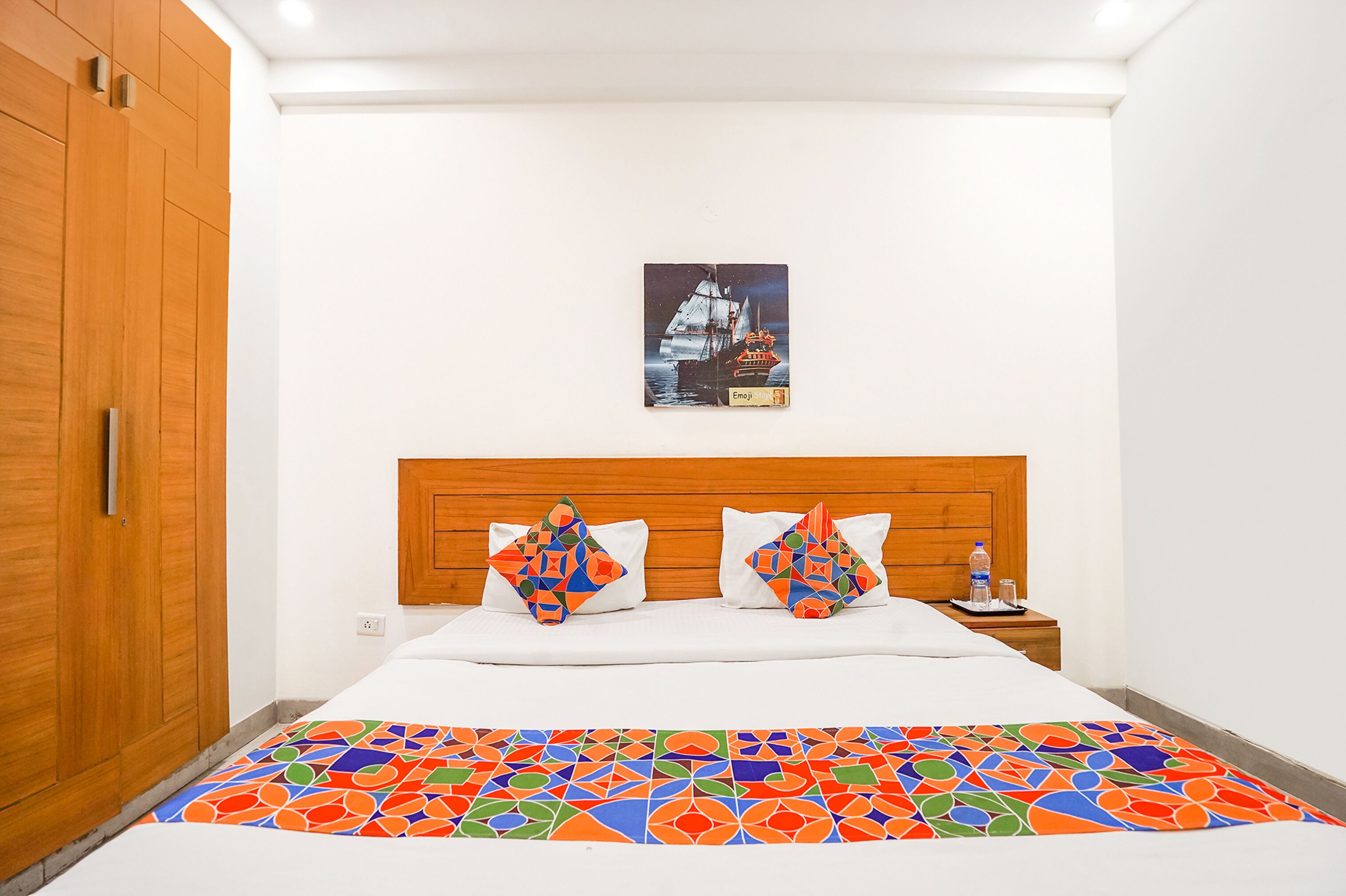 A bedroom with a wooden headboard, a bed with colorful geometric pillows, a framed picture of a ship on the wall, and a colorful geometric bedspread.