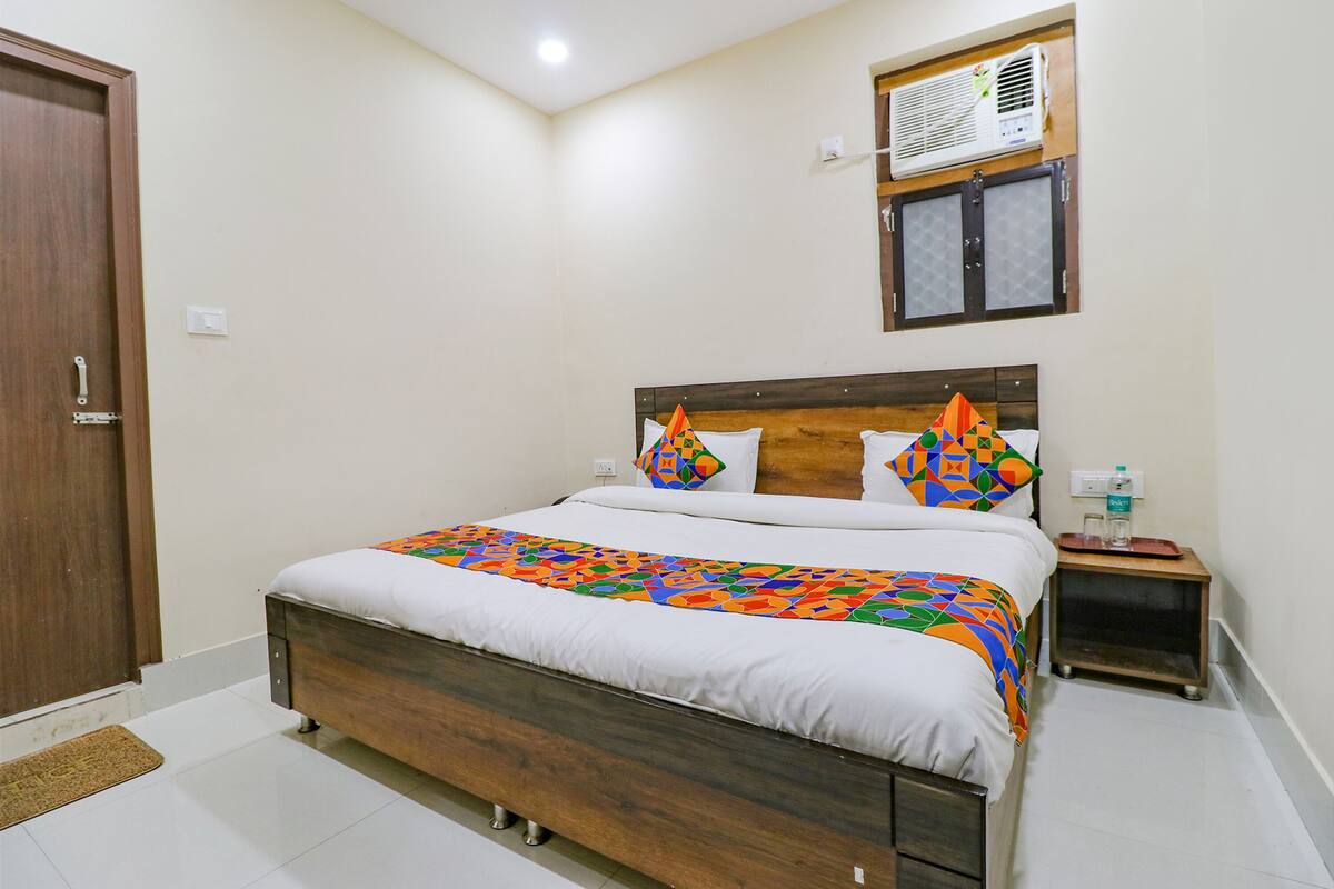 A bedroom with a wooden bed, white bedding, a colorful geometric throw, a small bedside table with a lamp, and a window with an air conditioner.