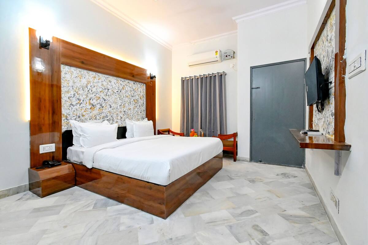 Un dormitorio con cabecera de madera, cama con ropa de cama blanca, mesita de noche con teléfono, cortina gris, televisor montado en la pared y puerta gris.