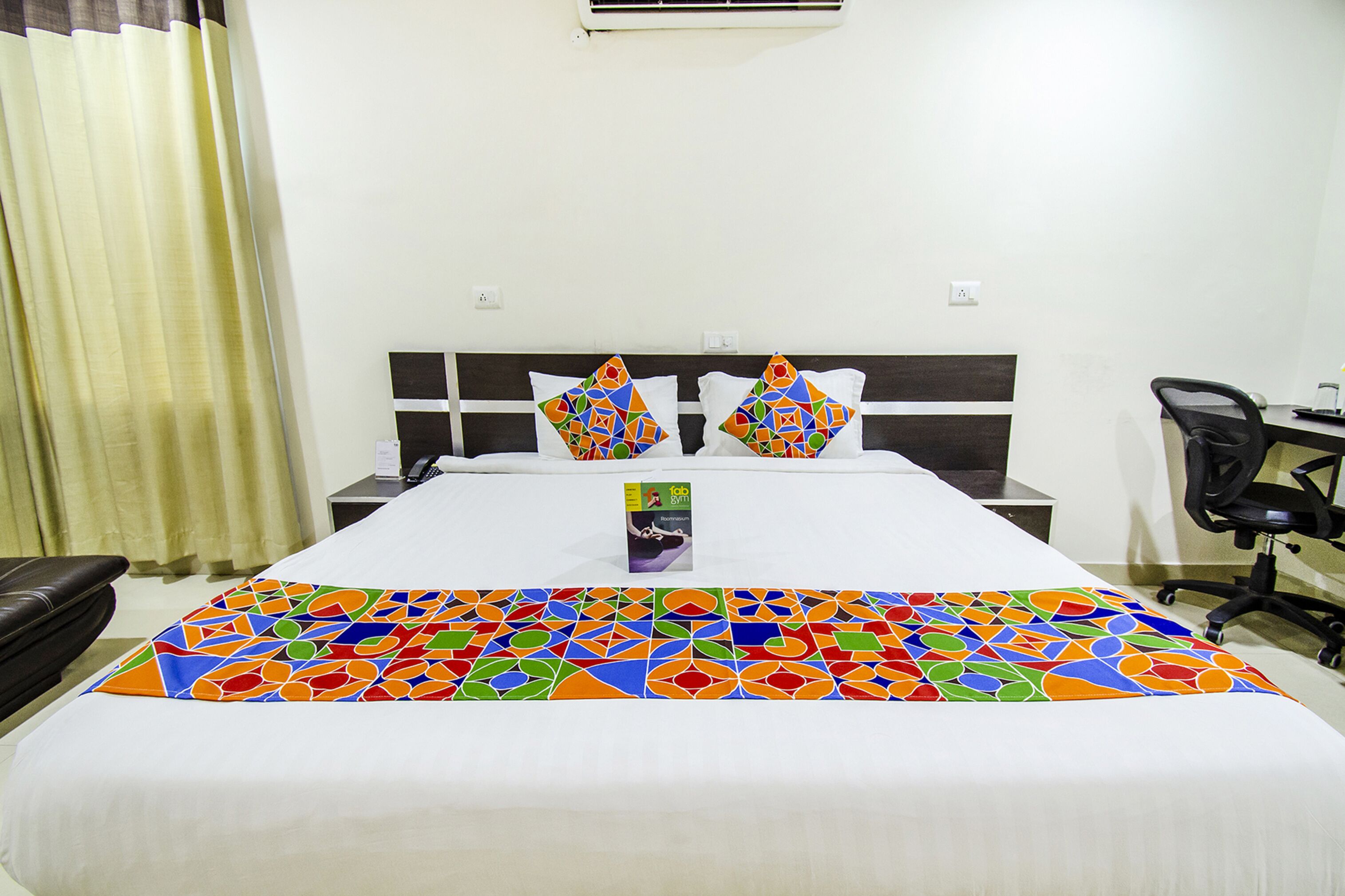Une chambre d’hôtel avec un lit bien fait, deux oreillers ornés d’un motif géométrique coloré, une table de chevet, une chaise et une petite table sur laquelle se trouve un livre.