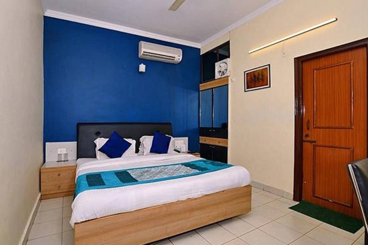 Un dormitorio con cama, mesita de noche, aire acondicionado instalado en la pared, puerta de madera y un cuadro enmarcado.