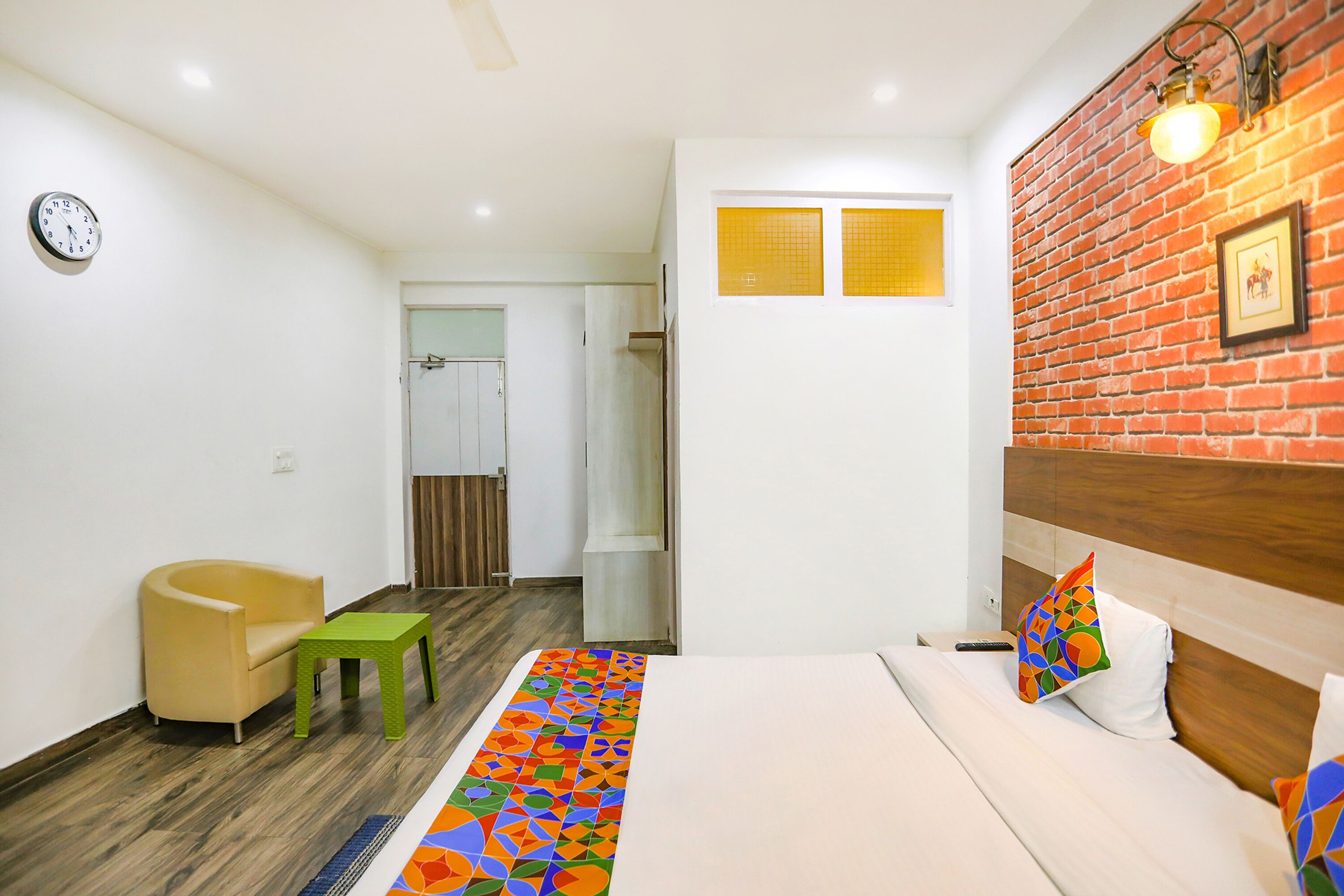 Quarto de hotel com cama, uma cadeira, uma mesinha, relógio e parede de tijolos.