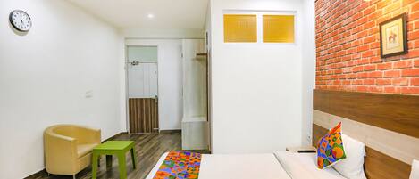 Quarto de hotel com cama, uma cadeira, uma mesinha, relógio e parede de tijolos.