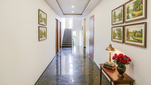 A hallway with a polished floor, framed landscape paintings on the walls, a staircase at the end, and a table with a lamp and flowers. - Fabhotel Shiv Rachna (Jaipur)