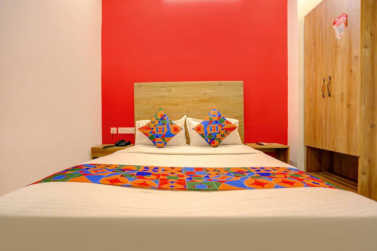 Une chambre d’hôtel avec une tête de lit en bois, un mur d’accent rouge, un lit recouvert d’une courtepointe aux motifs géométriques colorés, deux oreillers assortis et une armoire en bois.