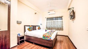 A hotel room with a wooden floor, a bed with a colorful bedspread, a bedside table, a wall-mounted air conditioner, and a window with patterned curtains. - FabHotel East Field Homes (Pune)