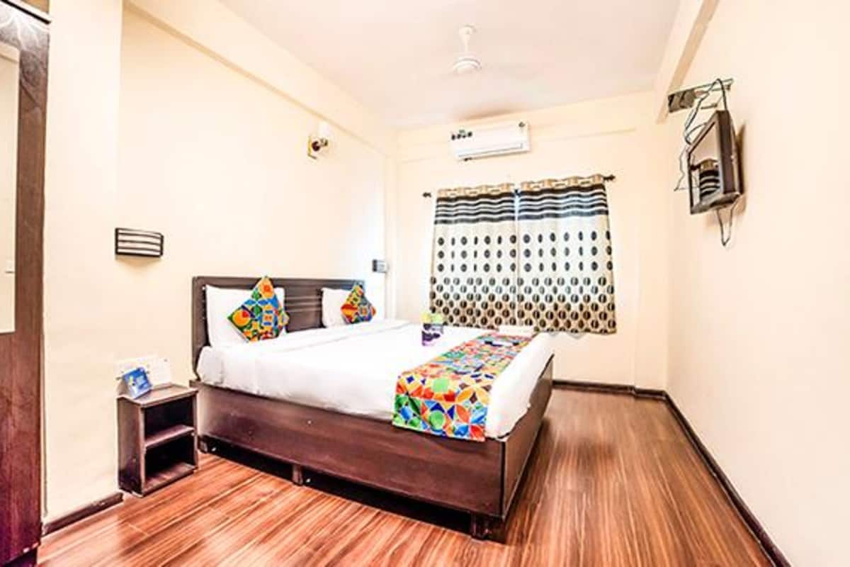 Una habitación de hotel con piso de madera, una cama con colcha colorida, una mesita de noche, un aire acondicionado instalado en la pared y una ventana con cortinas estampadas.