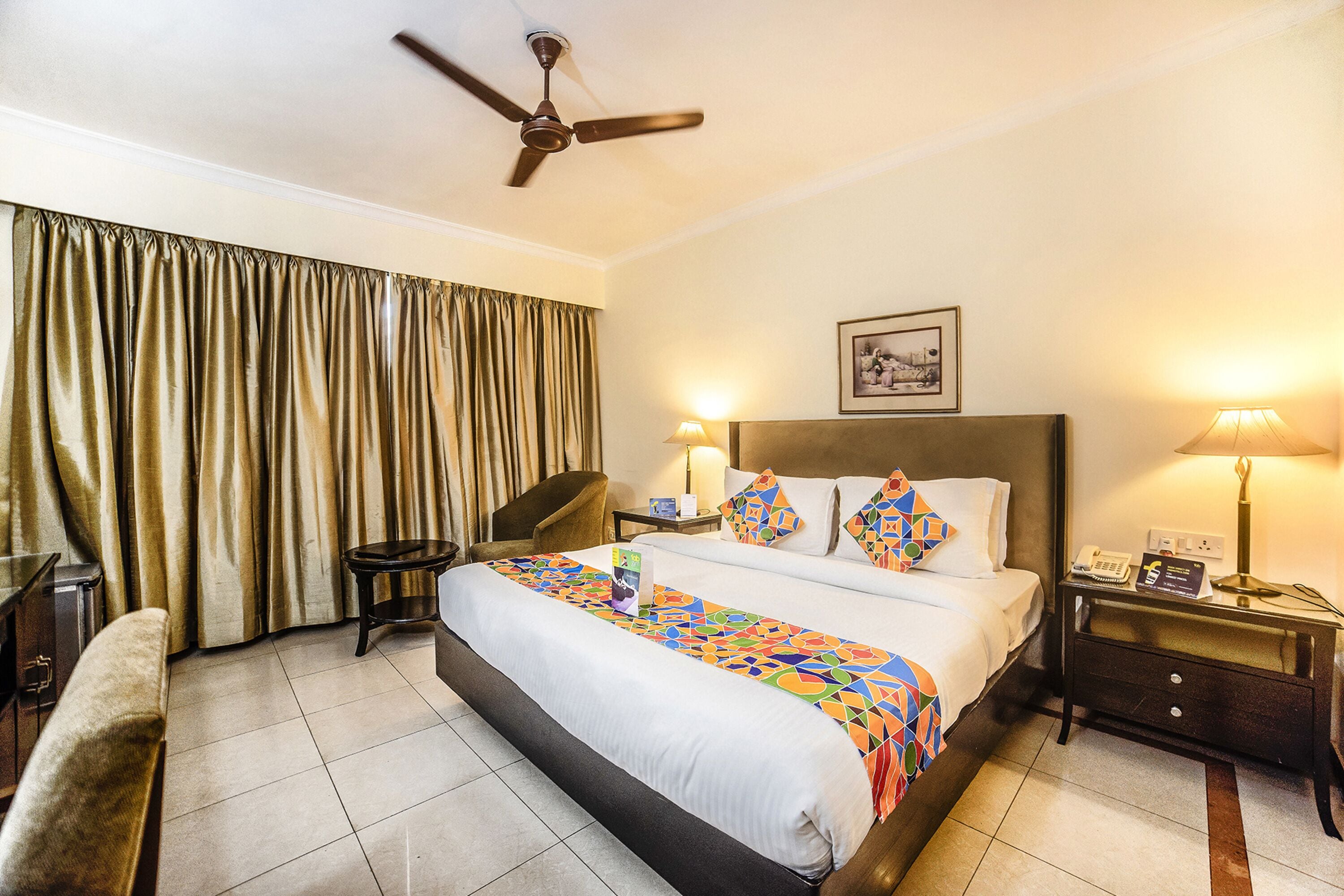 A hotel room with a large bed, a chair, a desk, a nightstand, a ceiling fan, and a window with curtains.