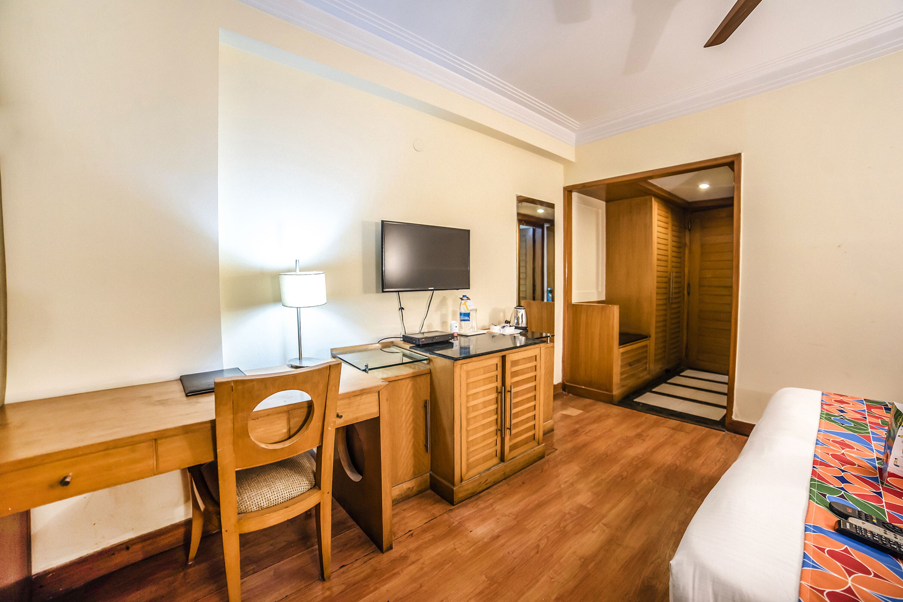 Habitación de hotel con un escritorio de madera, una silla, un televisor, una cama con colcha colorida y un armario de madera.