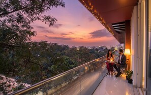 A balcony with a city view at sunset, a glass railing, and a table with two people.