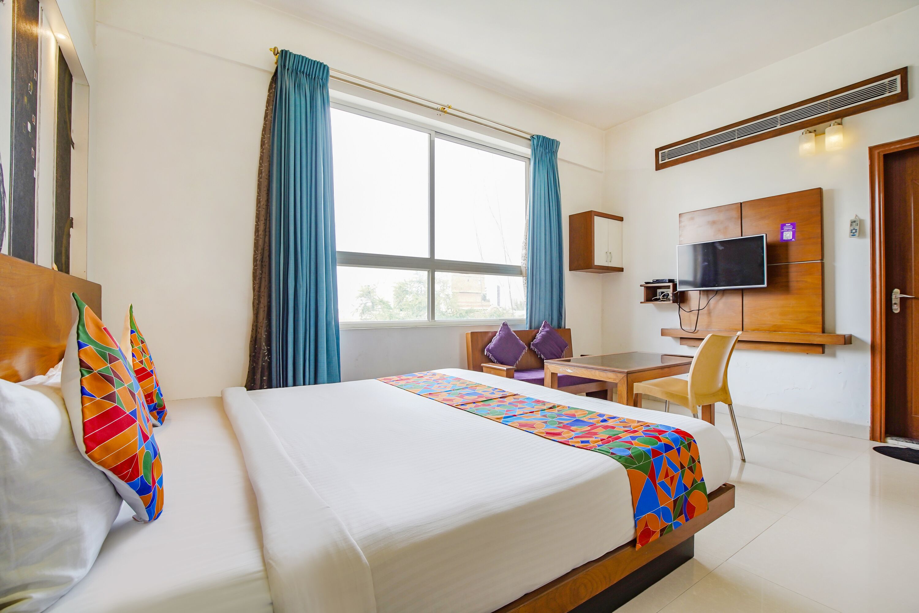 A hotel room with a large bed, a desk, a chair, a TV, and a window with curtains.