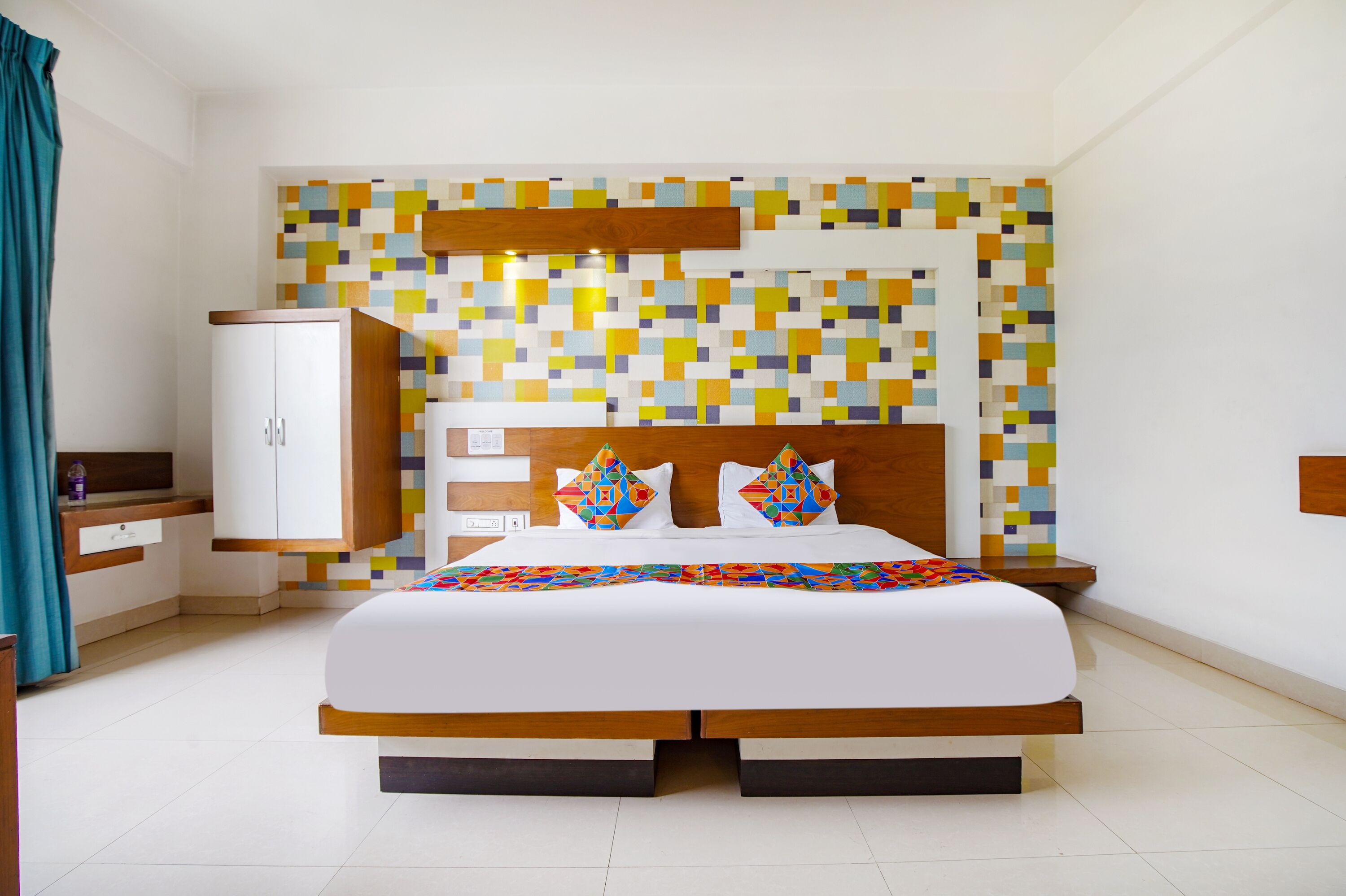A modern hotel room with a large bed, a wooden headboard, a colorful mosaic wall, and a white wardrobe.