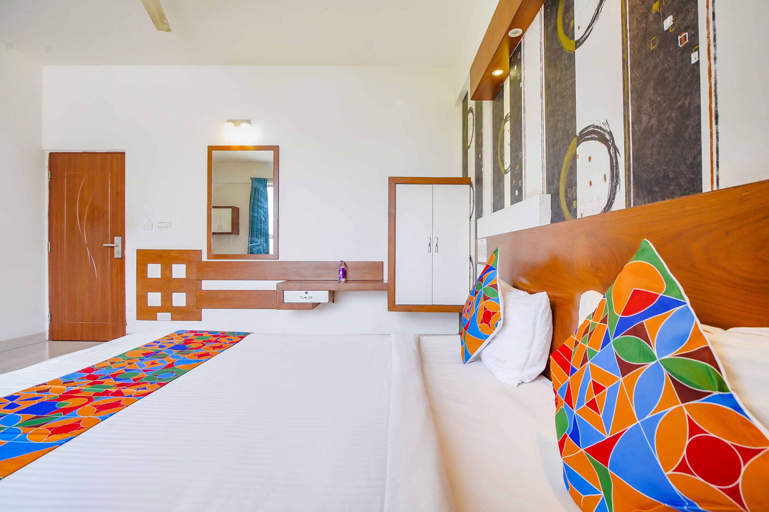 A hotel room with a bed, a wooden headboard, a colorful pillow, a white bedside table, a mirror, and a wardrobe.
