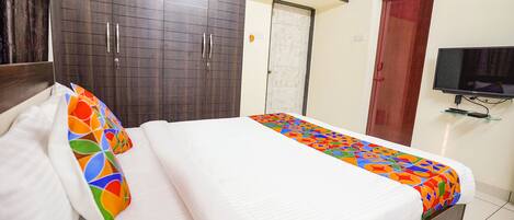 A neatly made bed with white linens and colorful pillows, a dark wooden wardrobe, a white door, and a mounted television on the wall.