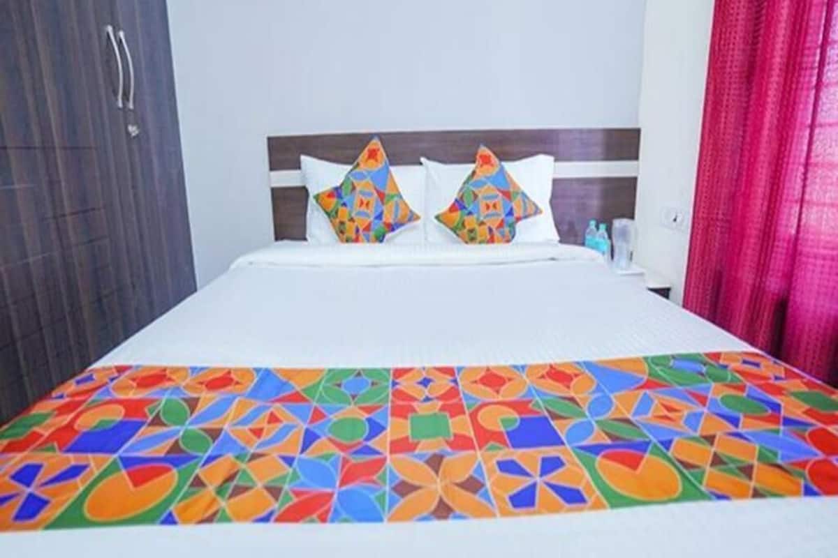 Un lit avec une couvre-lit aux motifs géométriques colorés, deux coussins décoratifs, une tête de lit en bois et un rideau rouge.