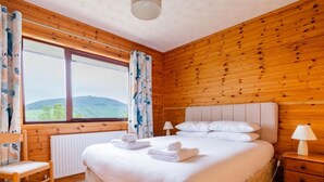 Une chambre avec un mur lambrissé en bois, un grand lit avec une literie blanche, une chaise, une table de chevet avec une lampe et une fenêtre avec des rideaux.
