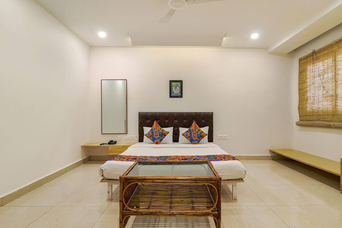 Une chambre d’hôtel comprenant un lit, une table de chevet, un miroir, une œuvre d’art accrochée au mur, une fenêtre avec des stores en bambou et un petit banc.