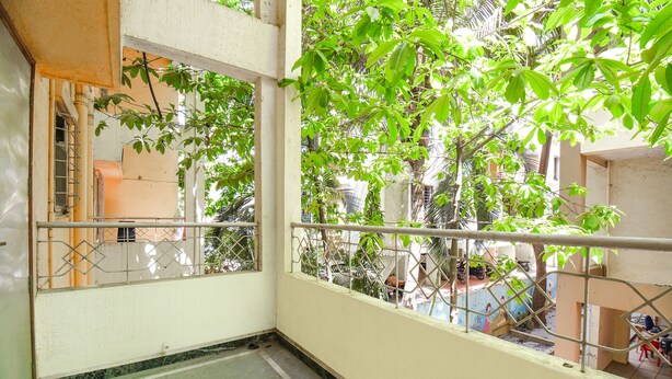 A balcony with a metal railing, a concrete floor, and a view of a residential area with trees and buildings. - FabExpress Wakad Stays (Paud)