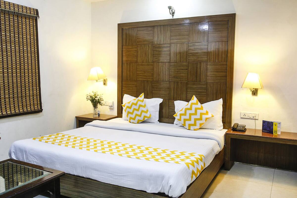 A hotel room with a large bed, two bedside tables, a nightstand, a lamp, and a small table with a vase.