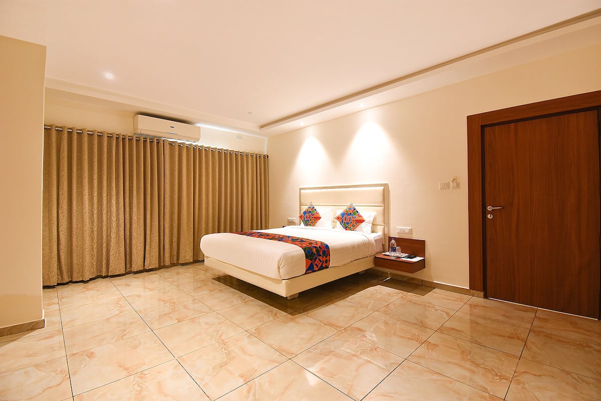 Quarto de hotel com cama, cortinas, unidade de ar condicionado, mesa de cabeceira e porta de madeira.