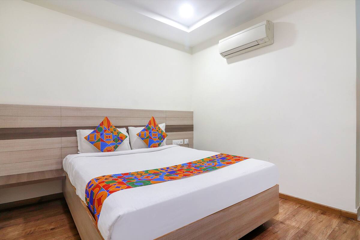 A neatly made bed with a colorful blanket, a wooden headboard, and a white wall with an air conditioning unit.