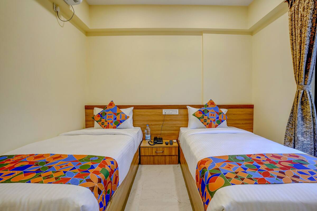 A hotel room with two beds, each with a wooden headboard, white bedding, and colorful geometric-patterned bedspreads. There is a wooden nightstand between the beds with a water bottle, phone, and remote control.