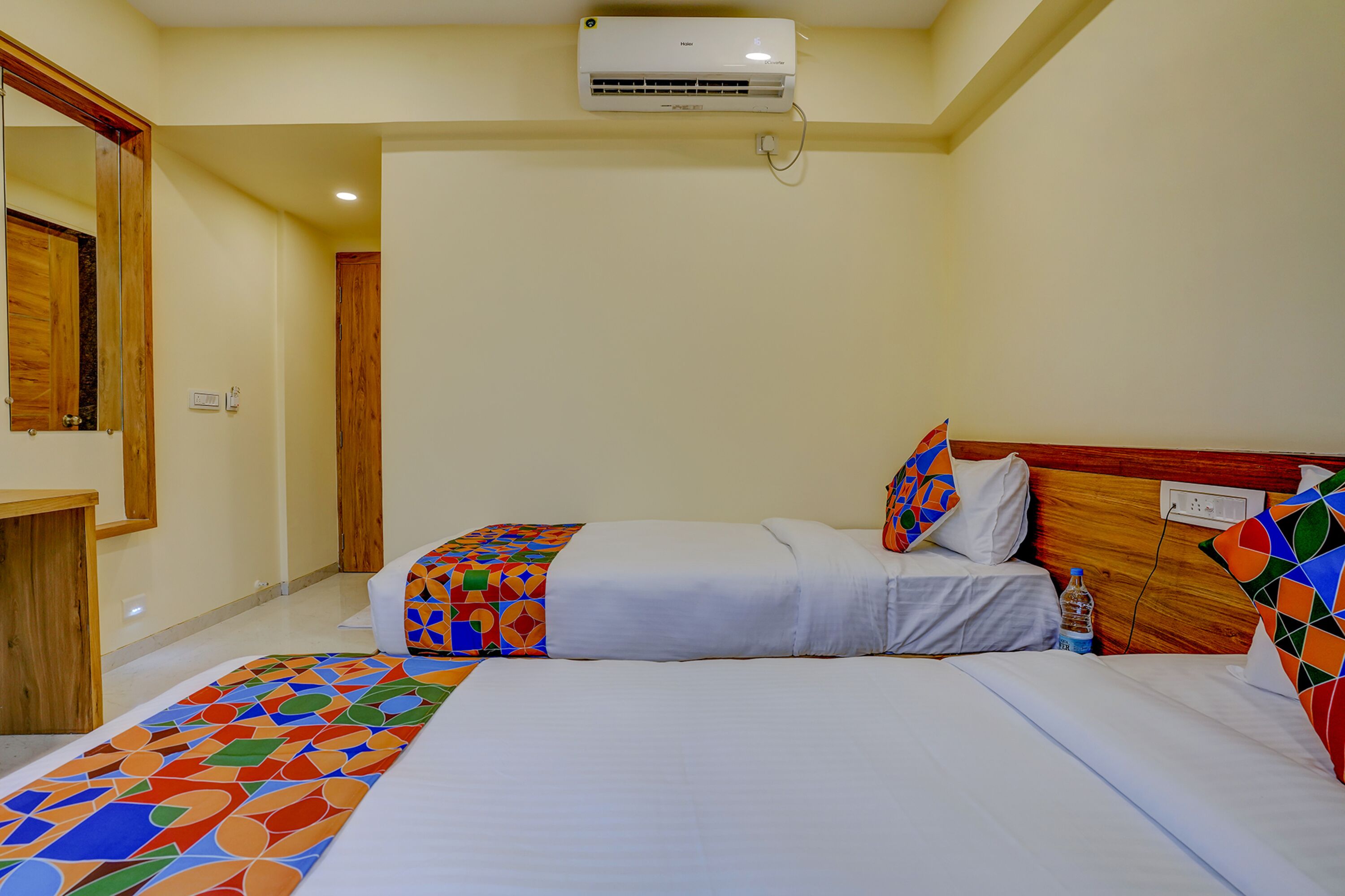 A hotel room with two beds, a wooden headboard, a colorful geometric-patterned bed runner, and a ceiling-mounted air conditioning unit.
