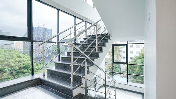 A modern staircase with metal railings, large windows, and a view of city buildings and greenery. - Fabhotel Crystal Homes (Pune)