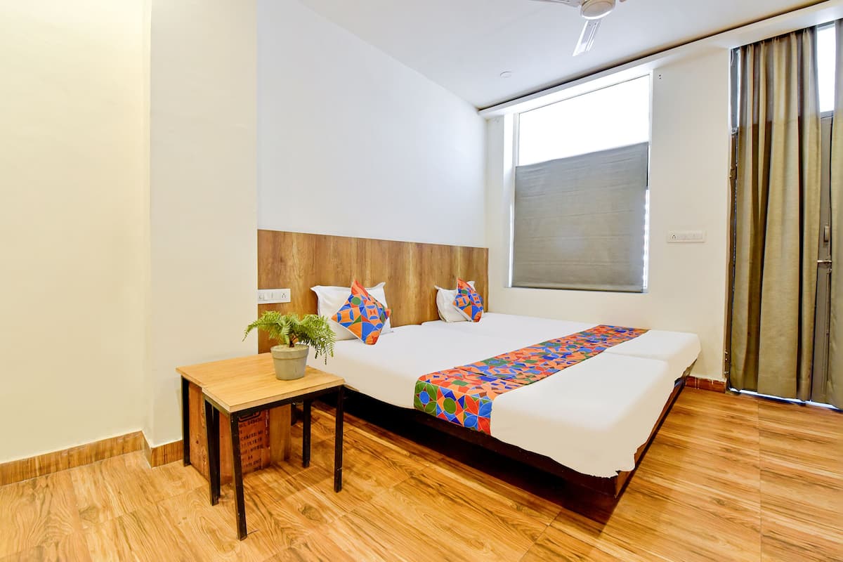 A hotel room with a wooden headboard, a large bed with a colorful geometric-patterned blanket, a small wooden table with a plant, and a large window with curtains.