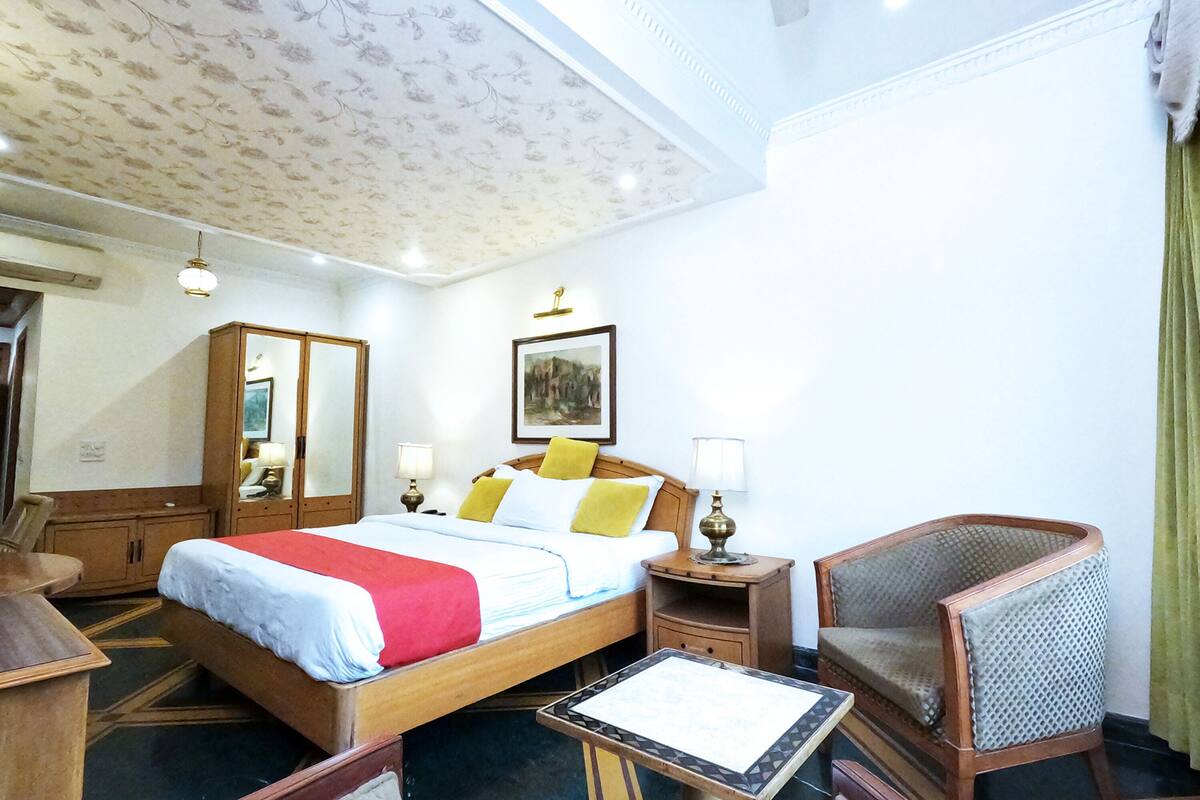 A hotel room with a bed, bedside table, chair, and a desk with a lamp.