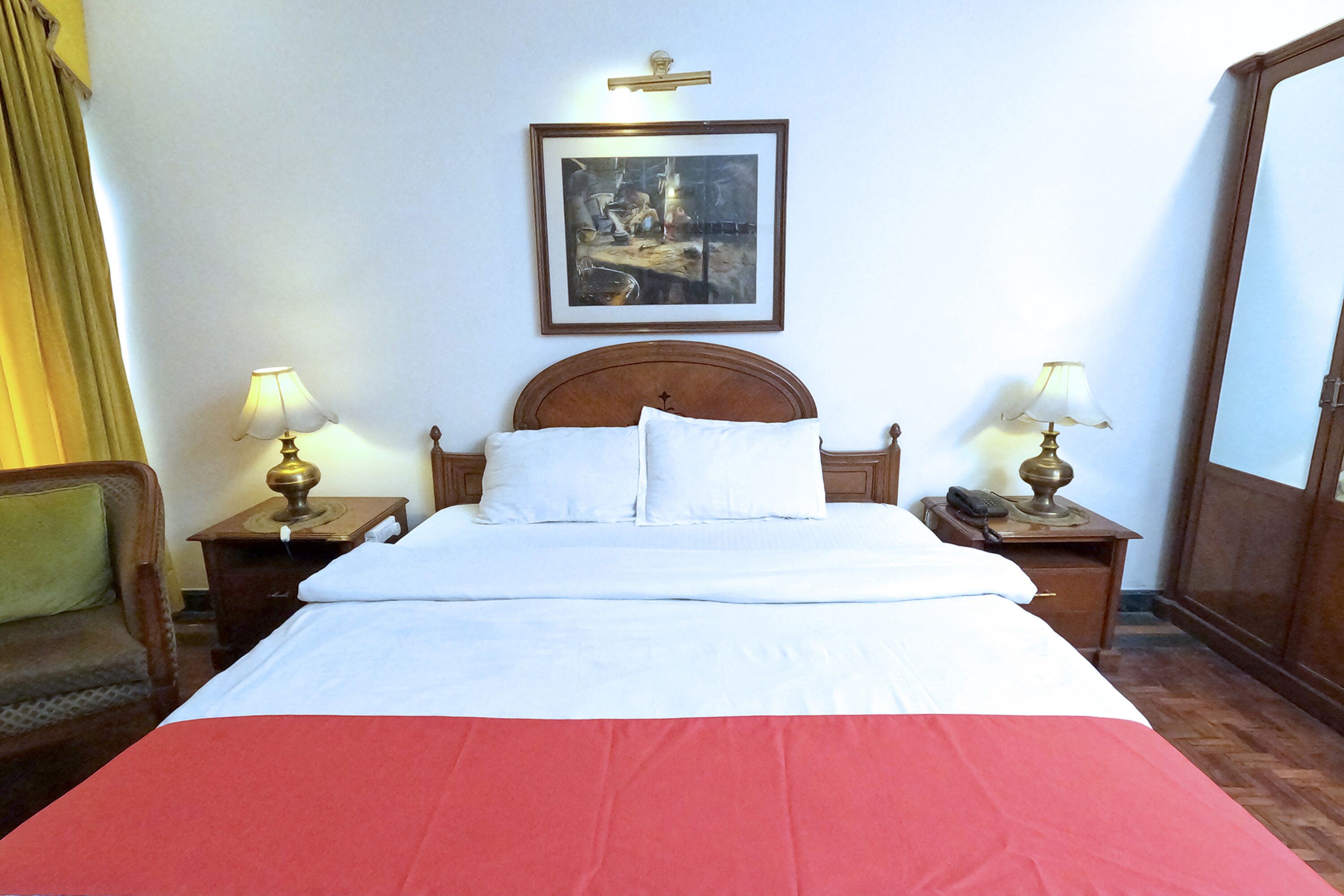 Cama bem arrumada com roupa de cama branca e vermelha, duas mesas de cabeceira com luminárias, um quadro na parede e uma porta de madeira.