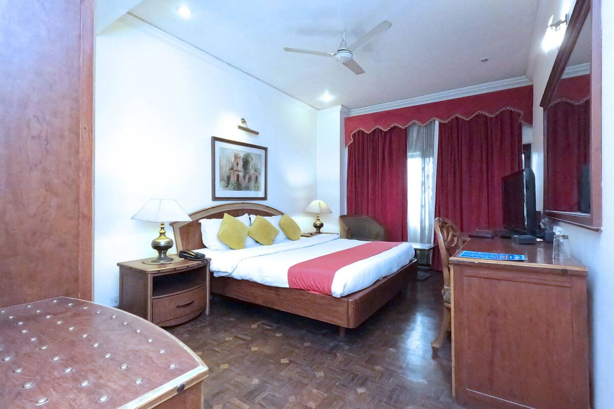 A hotel room with a wooden bed, a desk, a chair, a nightstand with lamps, a ceiling fan, and a framed picture on the wall.