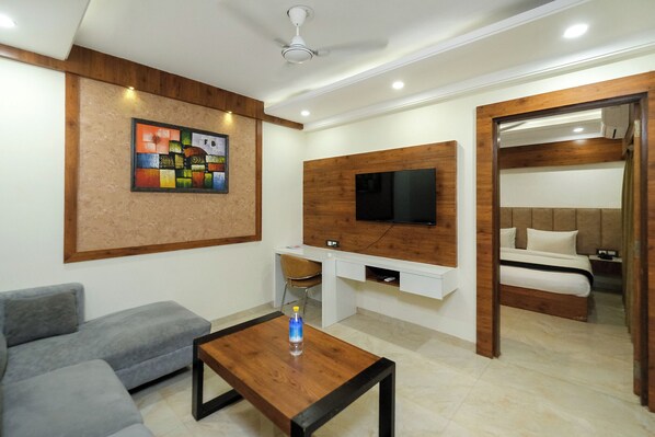 Uma sala de estar moderna com um sofá cinzento, uma mesa de centro de madeira, uma televisão de tela plana fixada em um painel de madeira e uma cama com cabeceira de madeira.