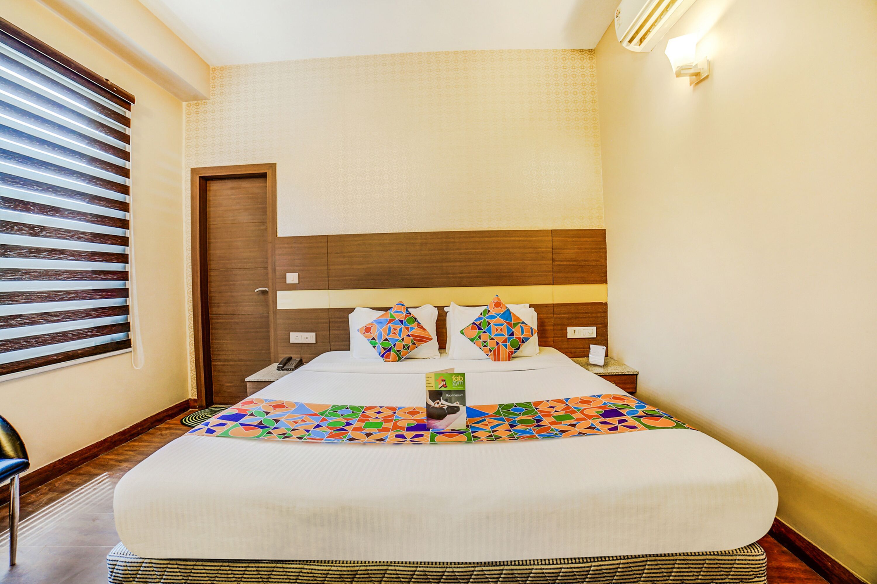Una habitación de hotel con una cama grande, un cabecero de madera, una mesita de noche con una lámpara, una ventana con persianas y una silla.