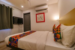 A hotel room with a bed, a television mounted on the wall, an air conditioning unit, and a framed picture on the wall.