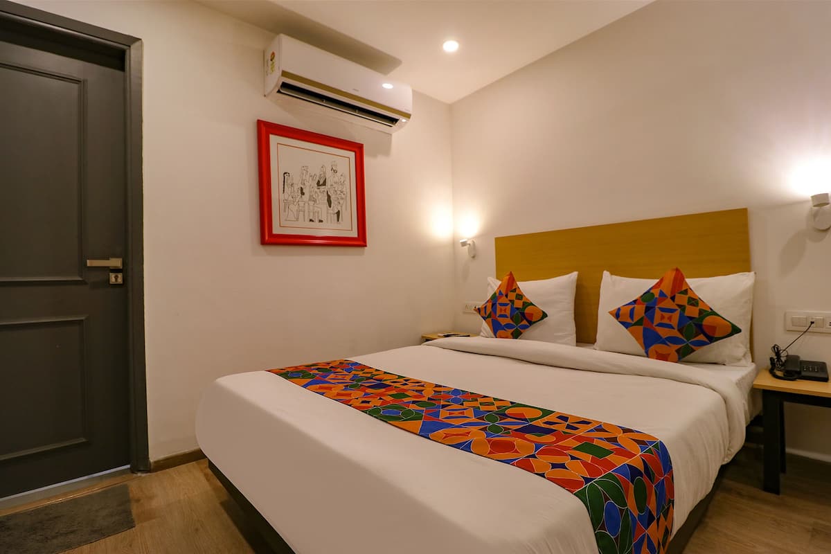 A hotel room with a bed, a nightstand, a wall-mounted air conditioner, and a framed picture on the wall.