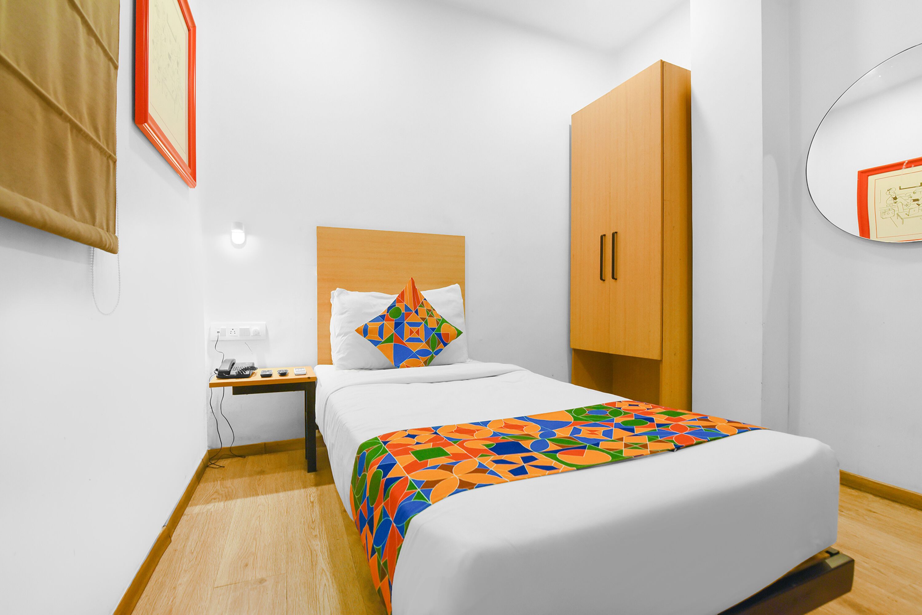 A neatly arranged hotel room with a bed, a wooden wardrobe, a bedside table with a lamp, and a wall-mounted mirror.